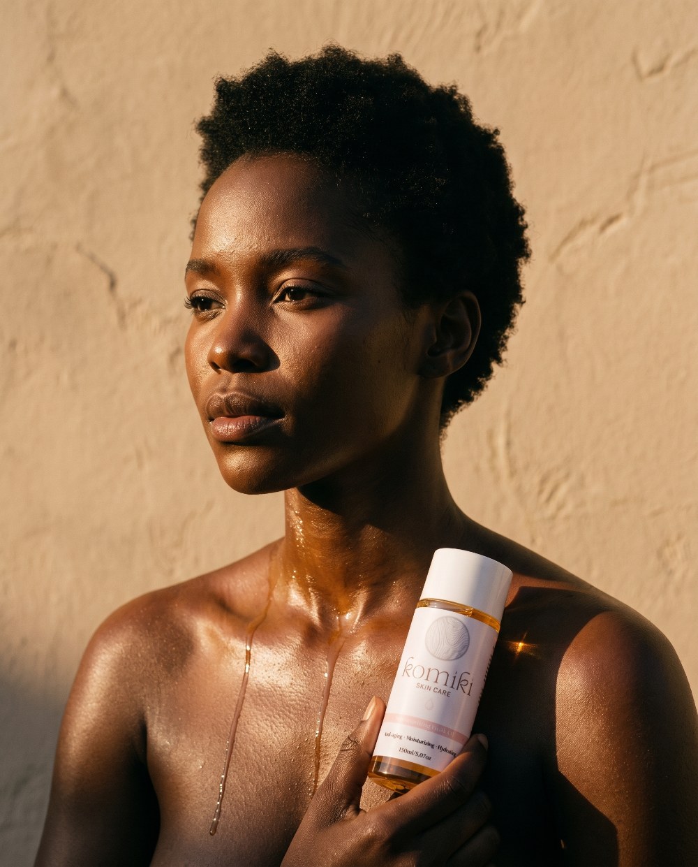 A woman in golden light, body oil glistening on her shoulders, holding Komiki Radiance Body Oil